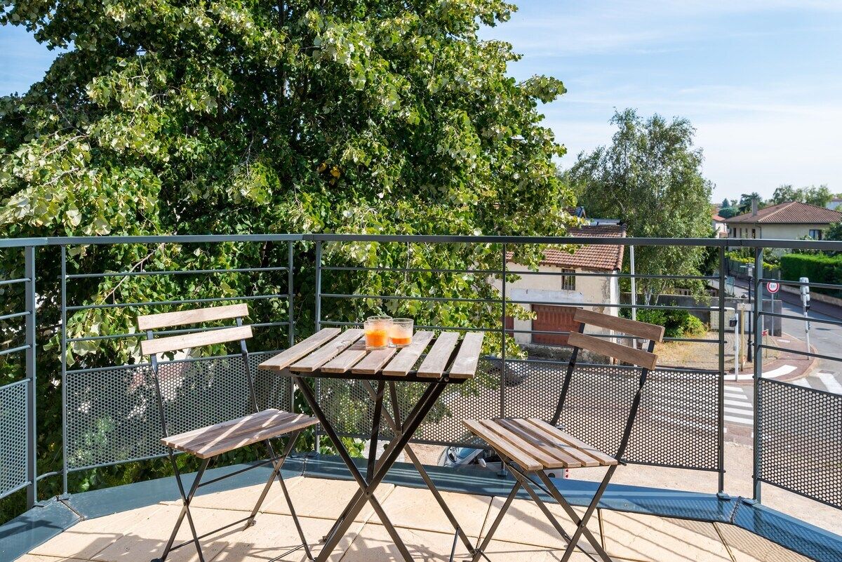 Un balcón con una mesa de madera y dos sillas, una verja de metal y un árbol grande al fondo.
