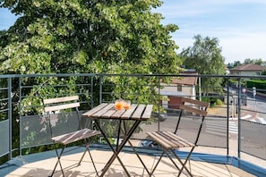 Un balcón con una mesa de madera y dos sillas, una verja de metal y un árbol grande al fondo.