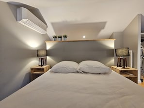 Un dormitorio con una cama, dos mesitas de noche, dos lámparas, un equipo de aire acondicionado y un estante con plantas.