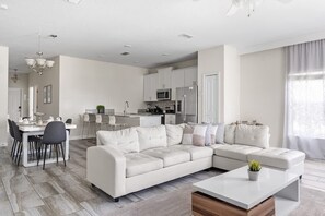 A modern living room with a sectional sofa, a dining area with chairs, a kitchen with cabinets and appliances, and a large window with curtains. - 5br/4ba Villa Sleeps 12 Private Pool & Spa (Kissimmee)