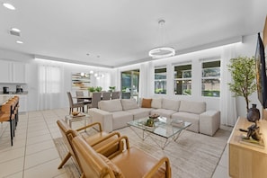 Ein modernes Wohnzimmer mit Sofa, Couchtisch und Sesseln. Es gibt einen Essbereich mit Tisch und Stühlen. Das Zimmer verfügt über große Fenster und einen Ausblick ins Freie.