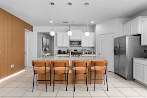 A modern kitchen with white cabinets, a stainless steel refrigerator, a microwave, and a bar area with four orange chairs. - Solterra Resort 6br/5.5ba Pool/spa Up to 18 (Davenport)