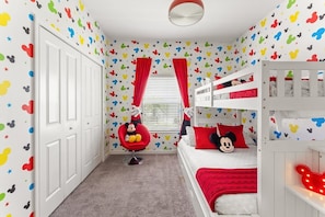 A child's room with Mickey Mouse-themed wallpaper, a bunk bed, a red chair, and a window with red curtains. - 5BR Home w/ Private Pool at Storey Lake (Kissimmee)