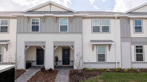 A two-story residential building with a symmetrical facade, a central entrance with double doors, and a small garden area with mulch and plants. - 5BR Themed Villa w/ Private Pool (Clermont)