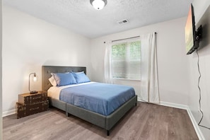 A bedroom with a bed, a nightstand with a lamp, a window with curtains, and a flat-screen TV mounted on the wall. - 3BR Woodstock | Pet Friendly | East Cobb Baseball (Woodstock)