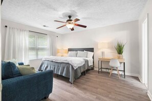 Une chambre à coucher comprenant un lit, un bureau, une chaise, un ventilateur de plafond et une fenêtre avec des rideaux.