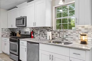 Una cocina moderna con armarios blancos, estufa de acero inoxidable, horno doble, lavavajillas, microondas, pileta con grifo moderno y ventana con vista a árboles.