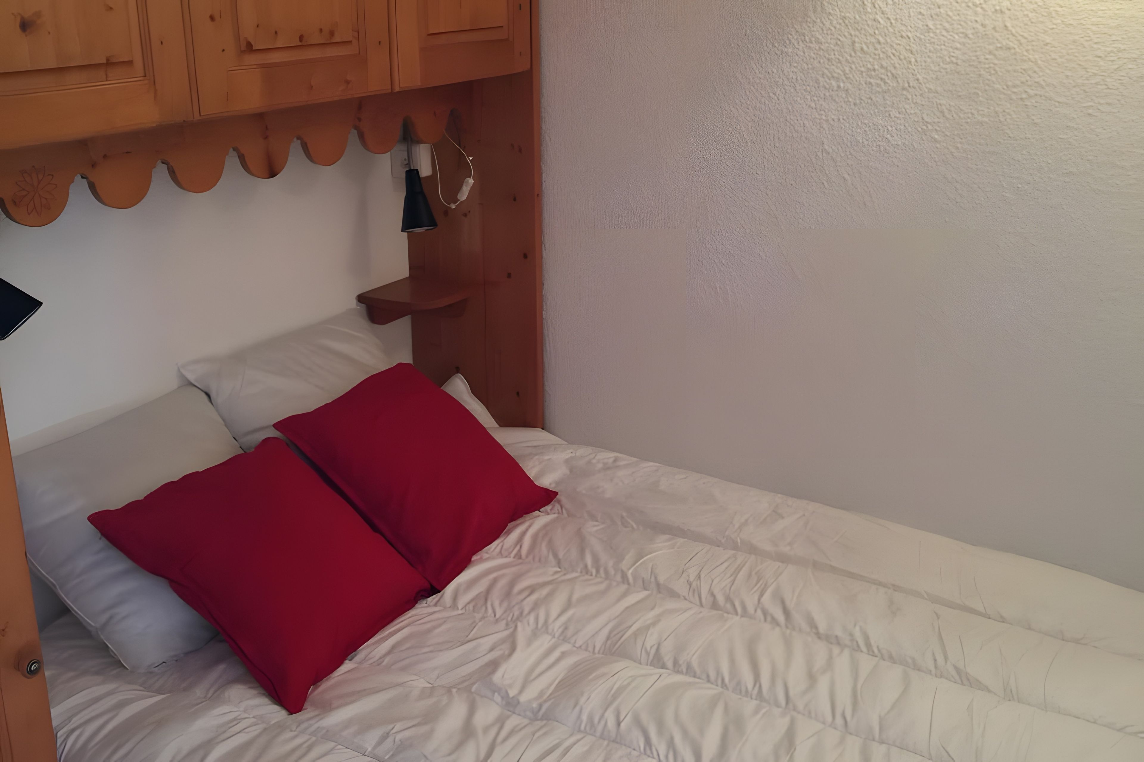 Ein Einzelbett mit weißer Bettwäsche und zwei roten Kissen, ein Holz-Kopfstück und eine kleine Ablage mit einer Lampe.