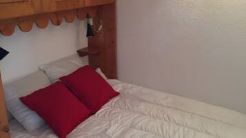 Ein Einzelbett mit weißer Bettwäsche und zwei roten Kissen, ein Holz-Kopfstück und eine kleine Ablage mit einer Lampe.