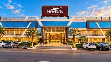 Ein Gebäude von Noventa Hotels & Resorts mit modernem architektonischen Design, das einen markanten Eingang und einen Parkplatz mit mehreren Autos zeigt.