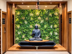 A statue of a seated figure in a meditative pose, surrounded by a lush green wall with hanging plants, set within a modern interior space with wooden paneling and recessed lighting.