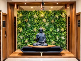 Una estatua de una figura sentada en posición meditativa, rodeada por un muro verde exuberante con plantas colgantes, ubicada en un espacio interior moderno con paneles de madera e iluminación empotrada.