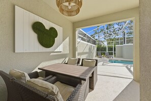 An outdoor patio with wicker furniture, a wooden table, and a pool area visible through a glass enclosure. - Luxury 4BR Townhome w/ Pool Champions Gate (Kissimmee)