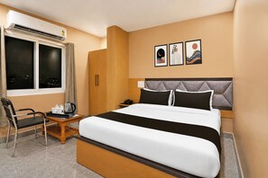 A hotel room with a bed, a desk with a chair, a window, and a wall with three framed pictures.