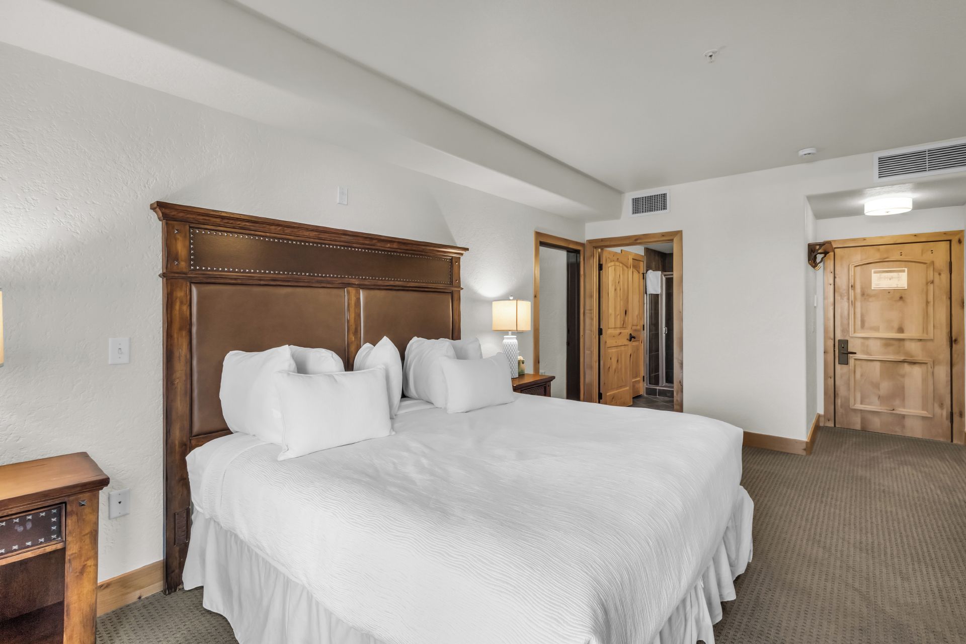 Une chambre d’hôtel avec un grand lit, une tête de lit en bois, une table de chevet, une lampe et une porte donnant accès à une salle de bain.