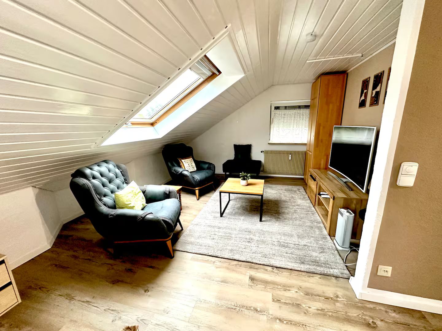 Ein Loft-Wohnzimmer mit schräg verlaufender Decke, einem schwarzen Ledersofa, einem Holztisch und einem Fernseher auf einem Holzständer.