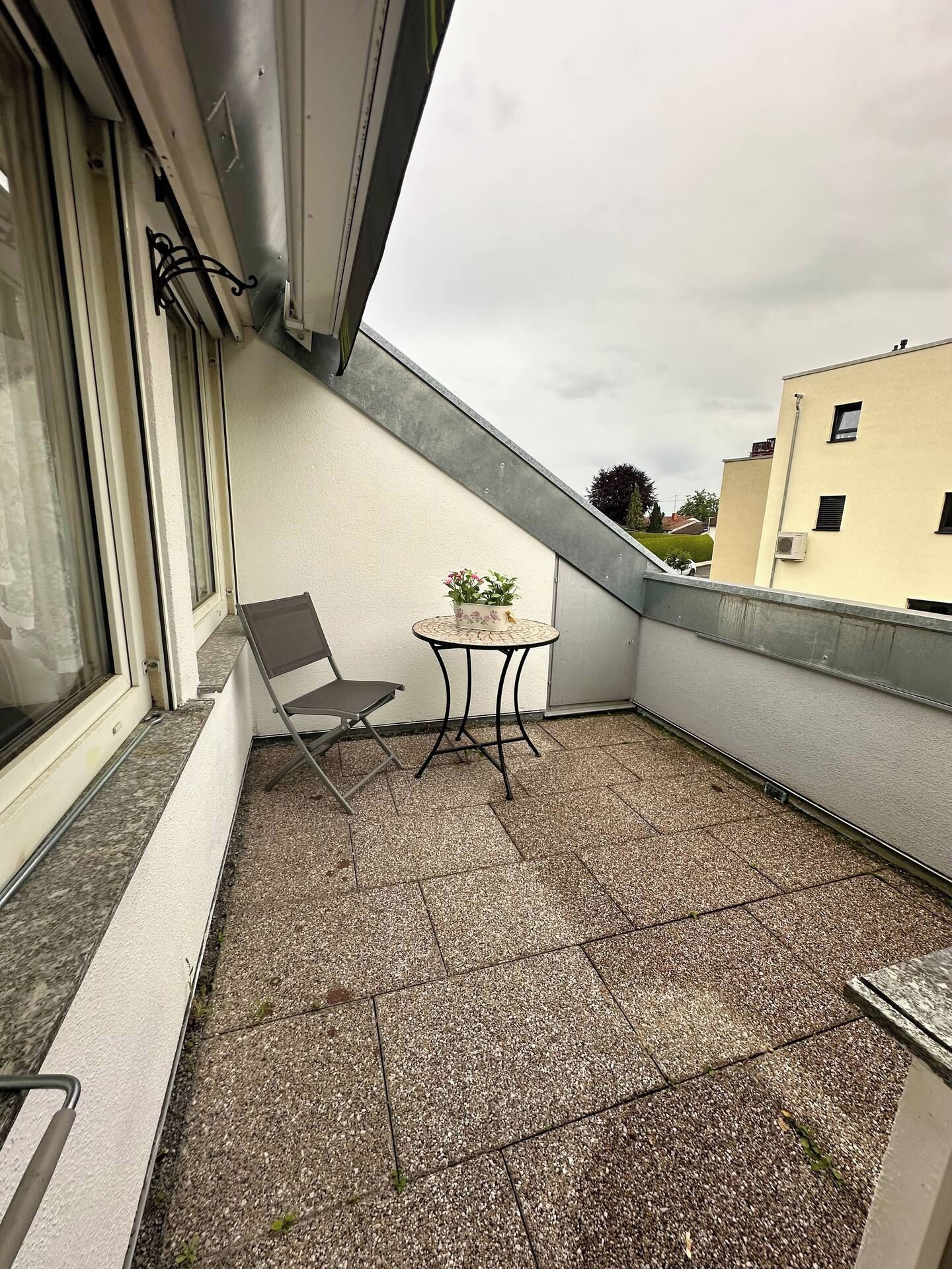 Ein kleiner Balkon mit Stuhl und Tisch, ein Fenster und Blick auf ein Gebäude.