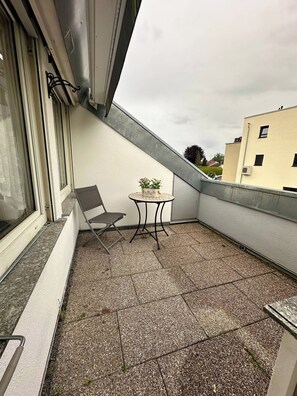 A small balcony with a chair and a table, a window, and a view of a building.