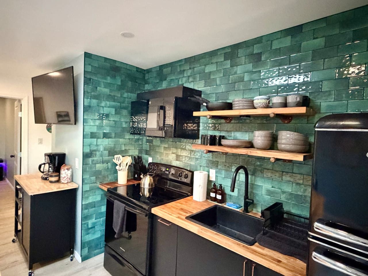 A compact kitchen with a black stove, a sink, and a microwave. There are shelves above the stove holding dishes.