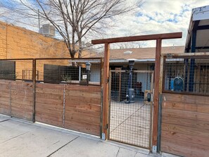 Un espace extérieur clos, délimité par une clôture en bois et une porte en bois, une clôture en métal et un bâtiment visible au loin.