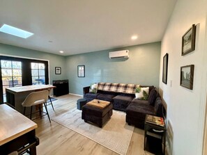 A living room with a sofa, ottoman, and a coffee table. There is a dining table with chairs, a small kitchen area, and a window with blinds.