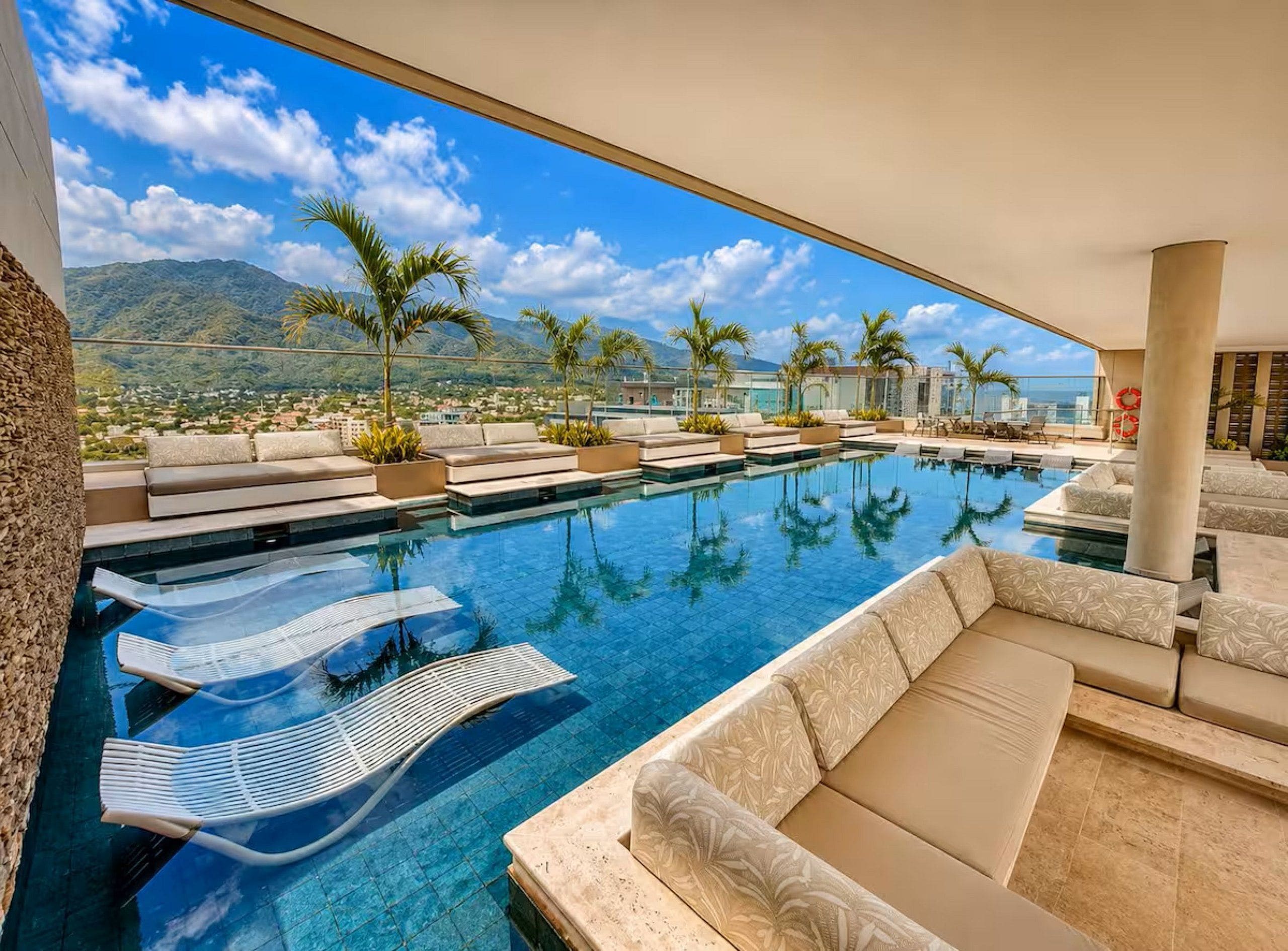 A rooftop infinity pool with lounge chairs and palm trees, overlooking a mountainous landscape.