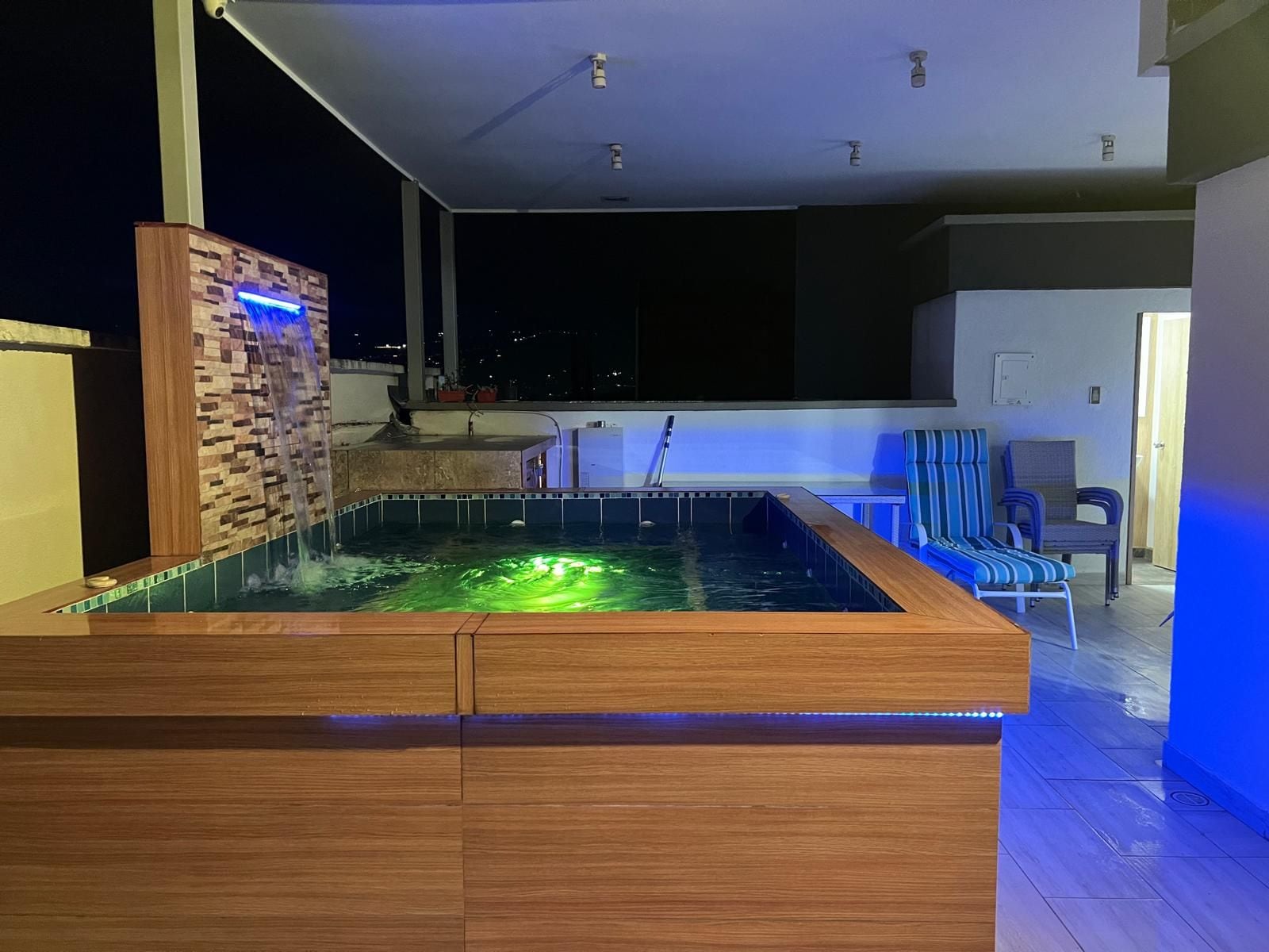 A modern outdoor hot tub with a waterfall feature, surrounded by a wooden deck and a seating area with chairs.