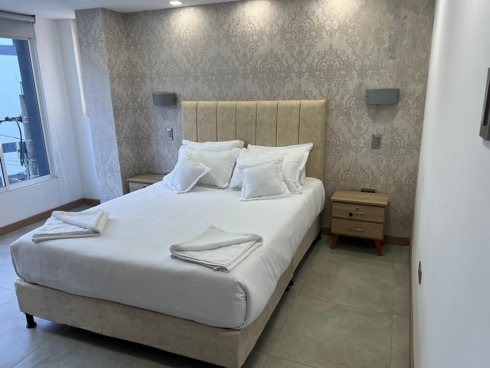 A neatly made bed with pillows and folded towels, a wooden nightstand with drawers, and a wall-mounted light fixture.