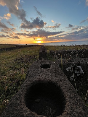 A sunset over a coastal landscape with ruins and a ship in the distance. - Hare Tupuna O Piri Nui (Hanga Roa)