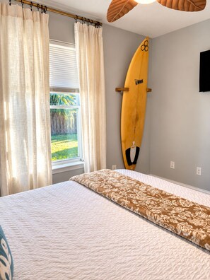 A bedroom with a bed, curtains, a surfboard, and a window with a view of greenery. - Endless Summer Surf Shack 4 min to beach Walk to Restaurants (Panama City Beach)