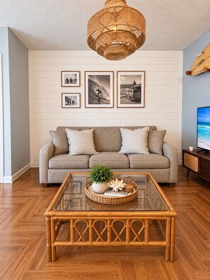 A living room with a sofa, a glass coffee table, and framed pictures on the wall. - Endless Summer Surf Shack 4 min to beach Walk to Restaurants (Panama City Beach)