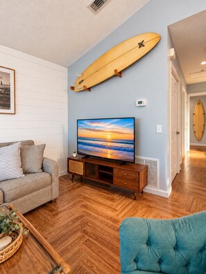 Un salon avec un plancher en bois, un canapé, un téléviseur et une planche de surf accrochée au mur.