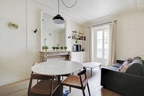 Un coin repas avec une table ronde et des chaises, un canapé, un téléviseur et une bibliothèque contenant des livres et des plantes.