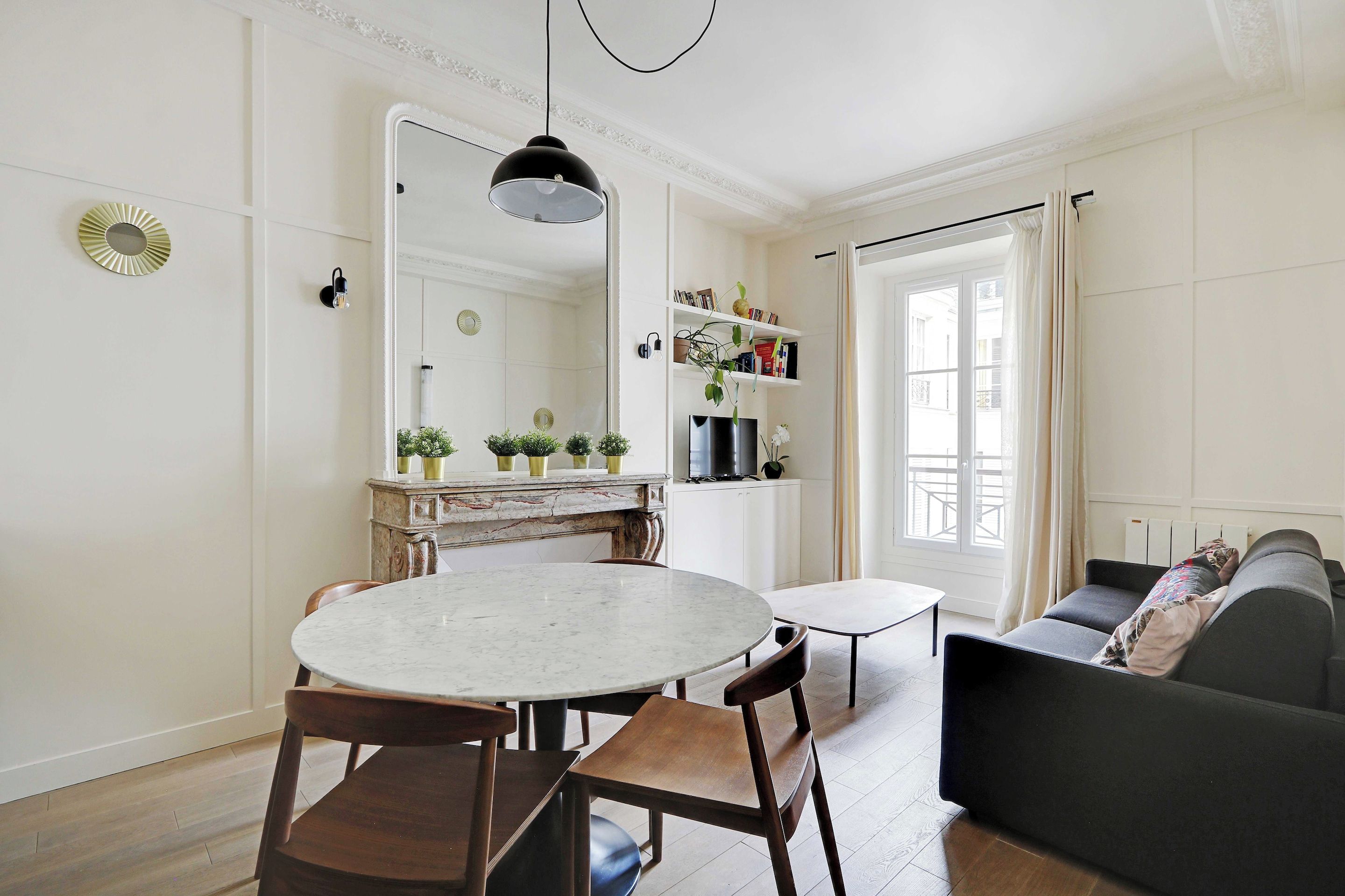 Un coin repas avec une table ronde et des chaises, un canapé, un téléviseur et une bibliothèque contenant des livres et des plantes.