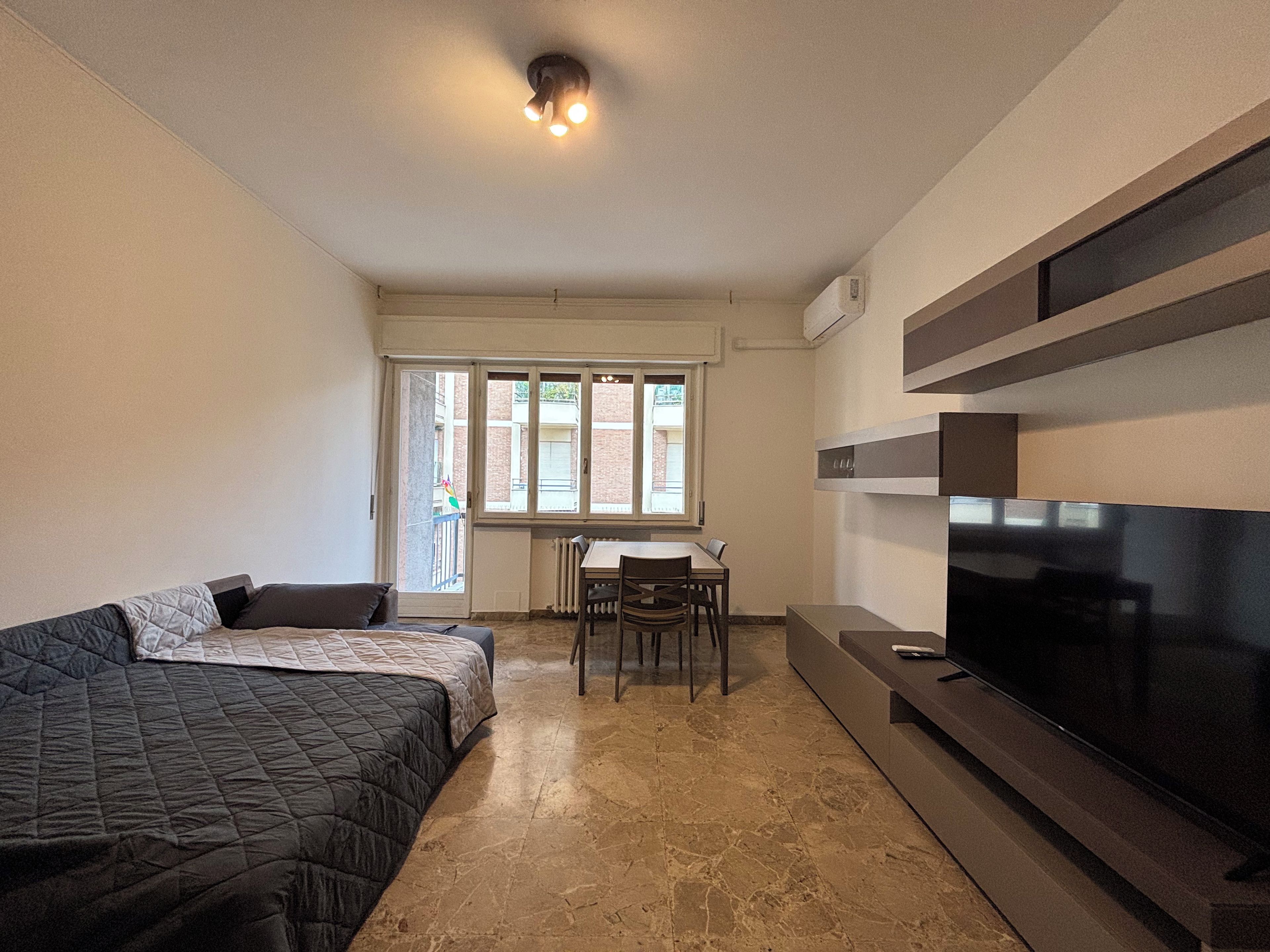 A bedroom with a bed, a TV, a dining table with chairs, and a window.