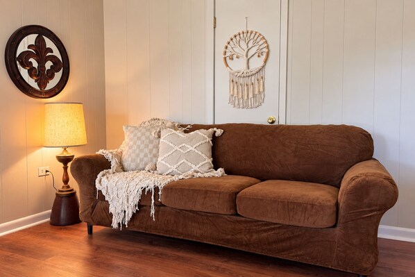 Ein braunes Sofa mit gemusterten Kissen und einer Wanddeko aus Makramee in einem Zimmer mit Holzboden und weißen Wänden.