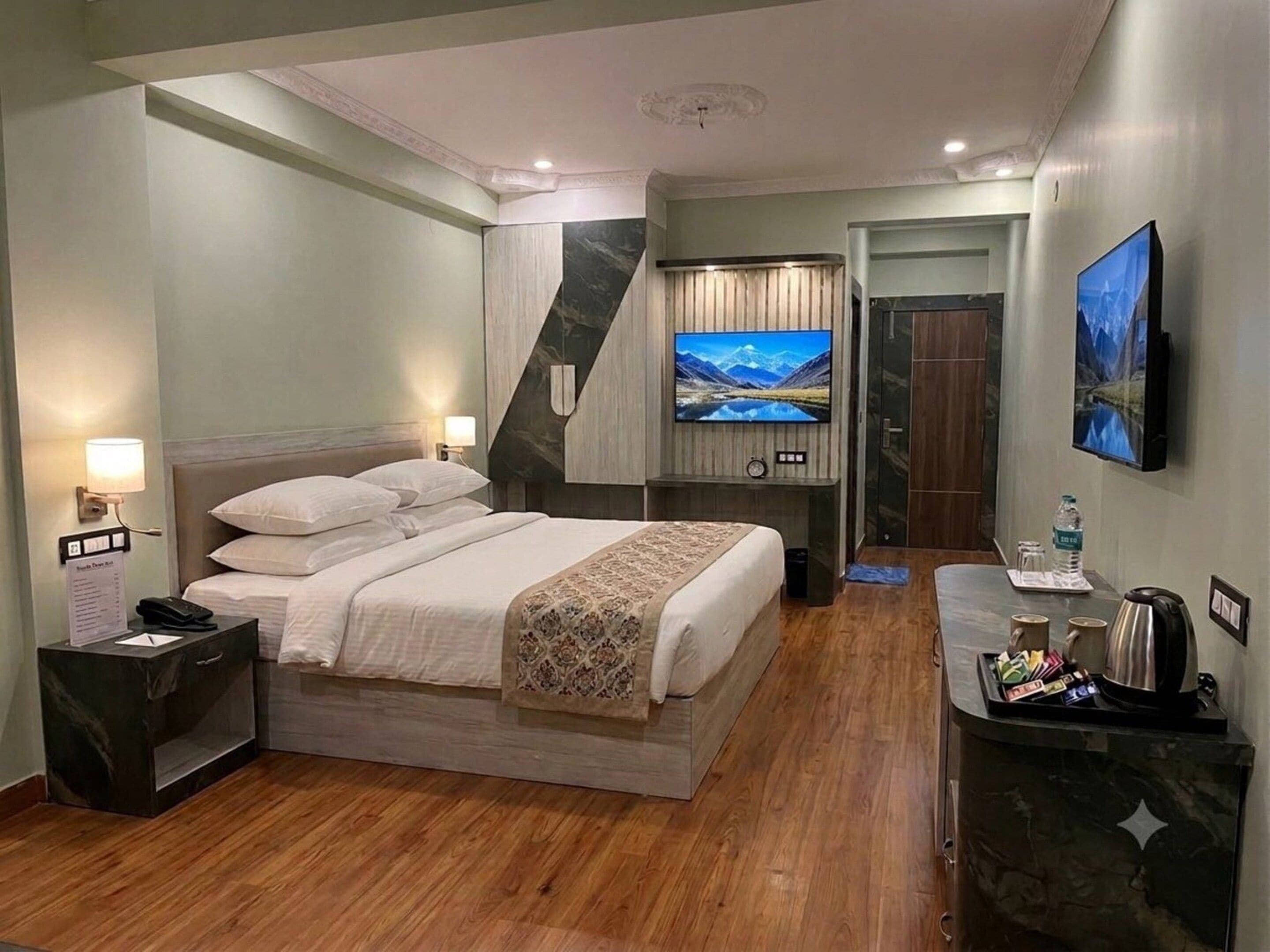 A hotel room with a bed, bedside table, lamp, TV, and a desk with a kettle and coffee cups.