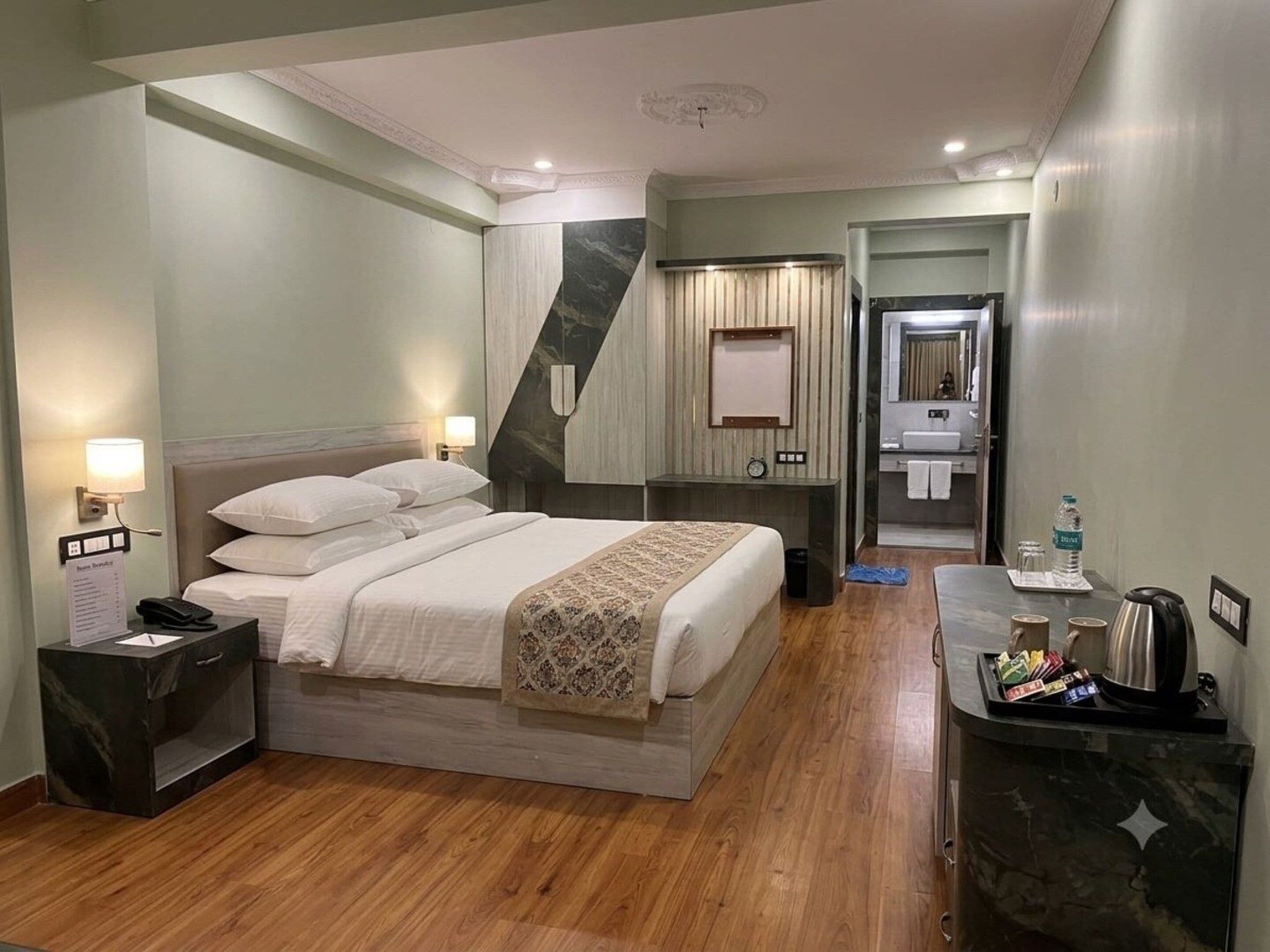 A hotel room with a bed, bedside table, lamp, and a desk with a kettle and coffee maker.