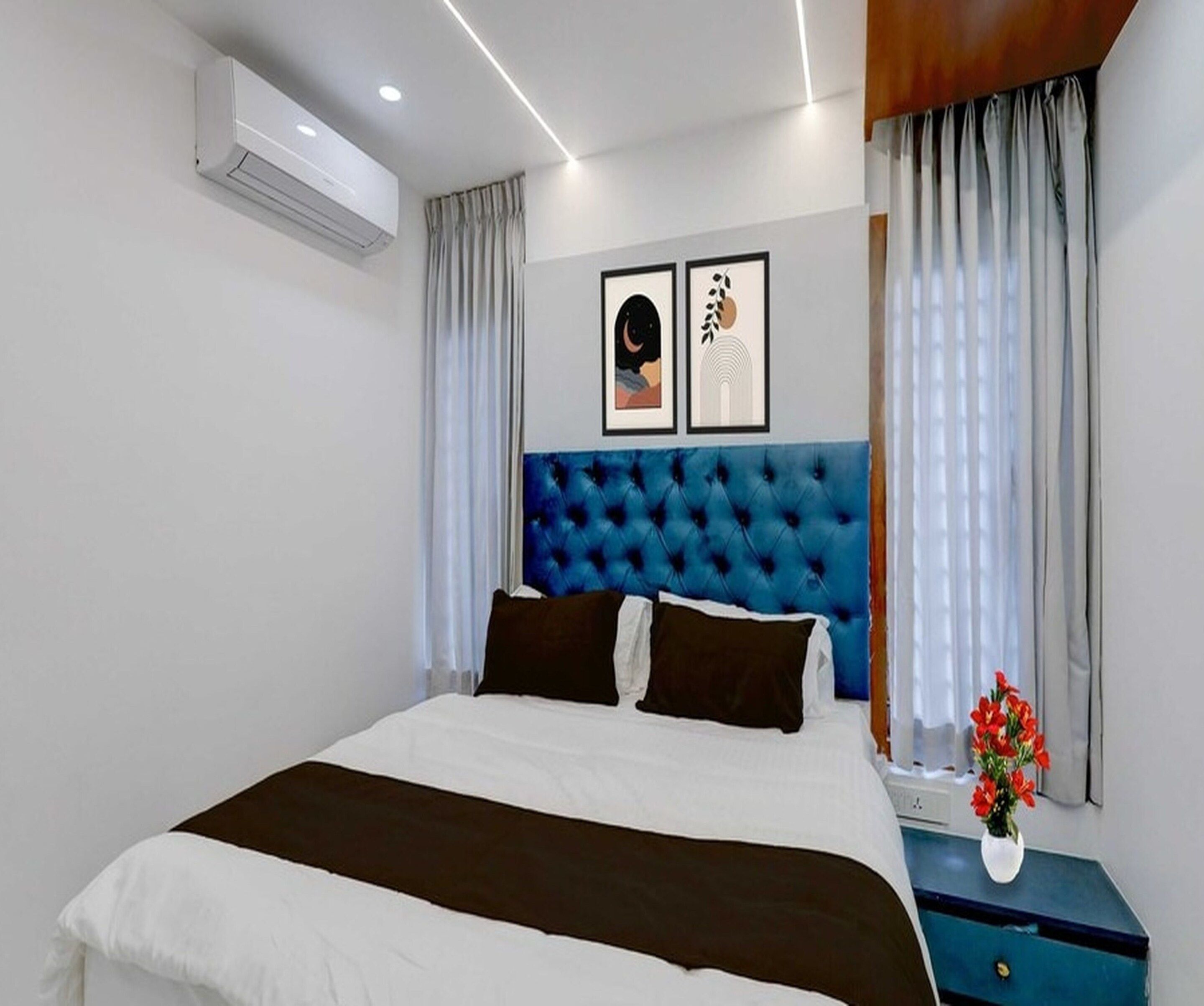 A hotel room with a large bed, a blue headboard, two framed pictures on the wall, and a nightstand with a vase of flowers.
