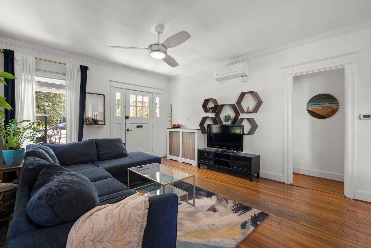 Un salon avec un canapé, une table basse et un meuble pour la télévision. On y trouve également un ventilateur de plafond, une porte et une fenêtre avec des rideaux.