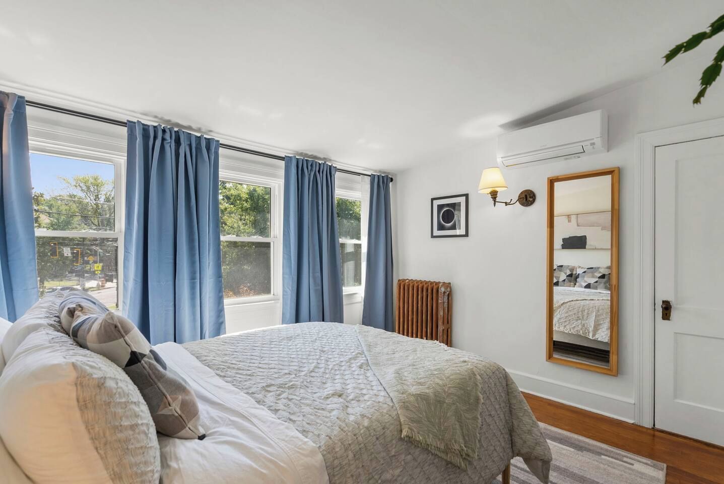 A bedroom with a bed, a radiator, a window with curtains, a wall-mounted lamp, and a mirror.