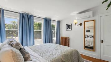 A bedroom with a bed, a radiator, a window with curtains, a wall-mounted lamp, and a mirror.