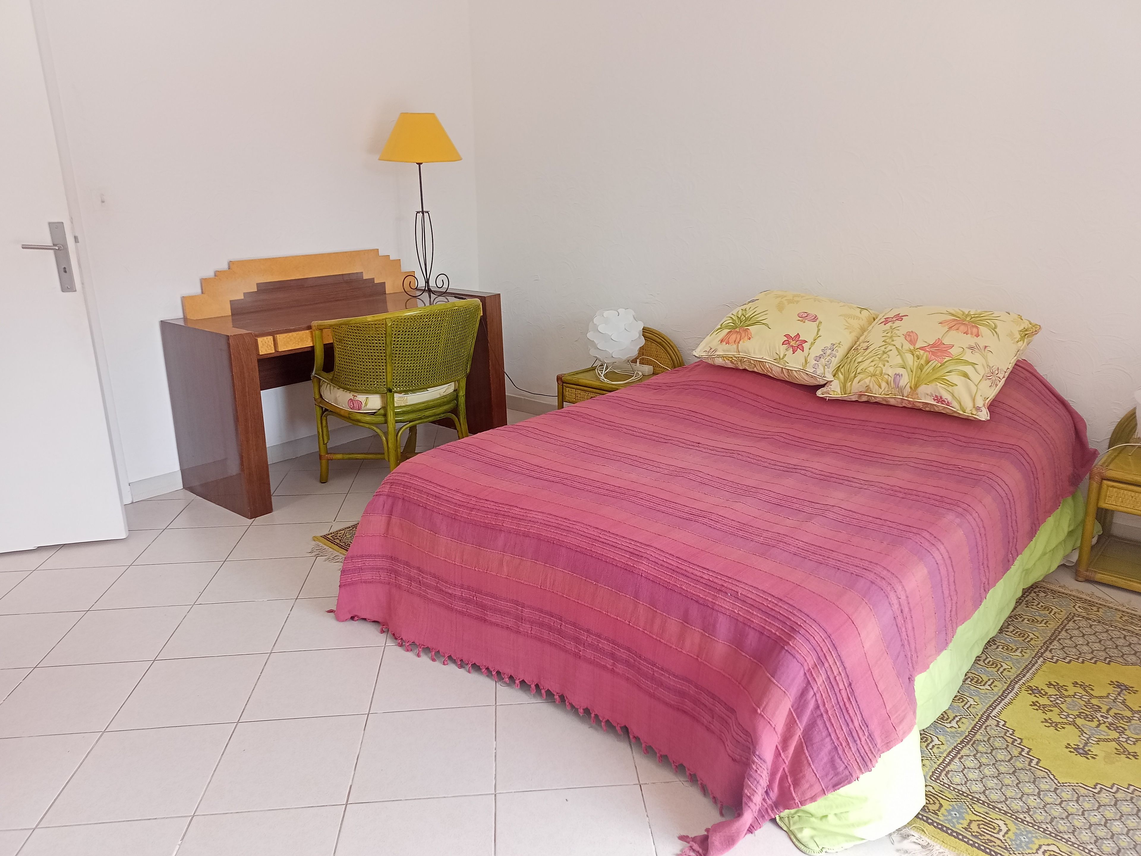 Een slaapkamer met een bed, een bureau, een stoel, een lamp en een nachtkastje.