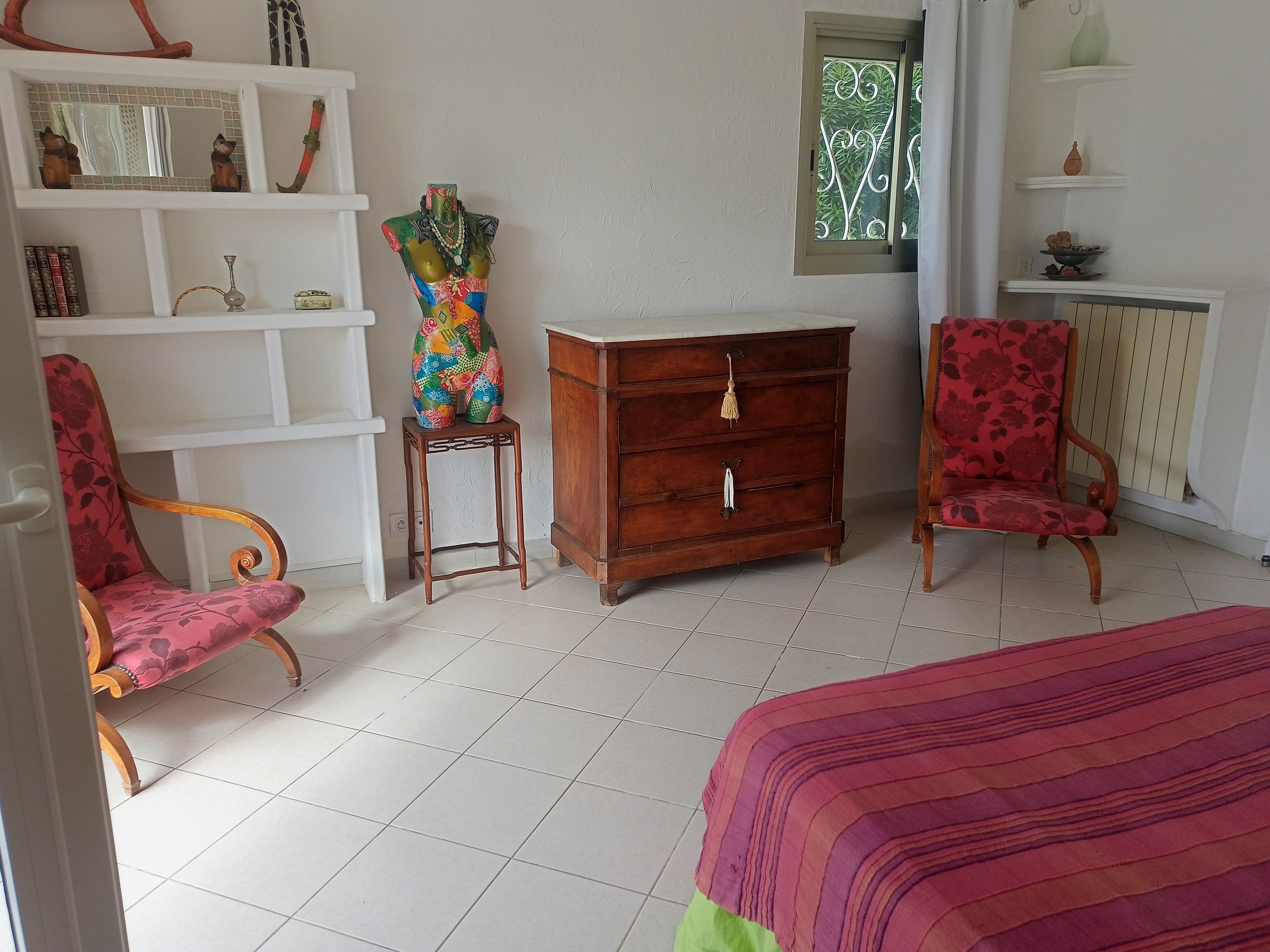Una habitación con cama, dos sillas, cómoda, estante con adornos y ventana con vistas.