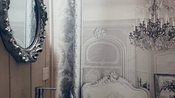 Um banheiro com um espelho grande e ricamente decorado, pia em vidro e uma cadeira clássica.