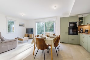 A modern living room with a sofa, dining table, and TV. There is a kitchen area with cabinets and appliances. - Quartz - Jardin - Proche plage (Saint-Hilaire-de-Riez)