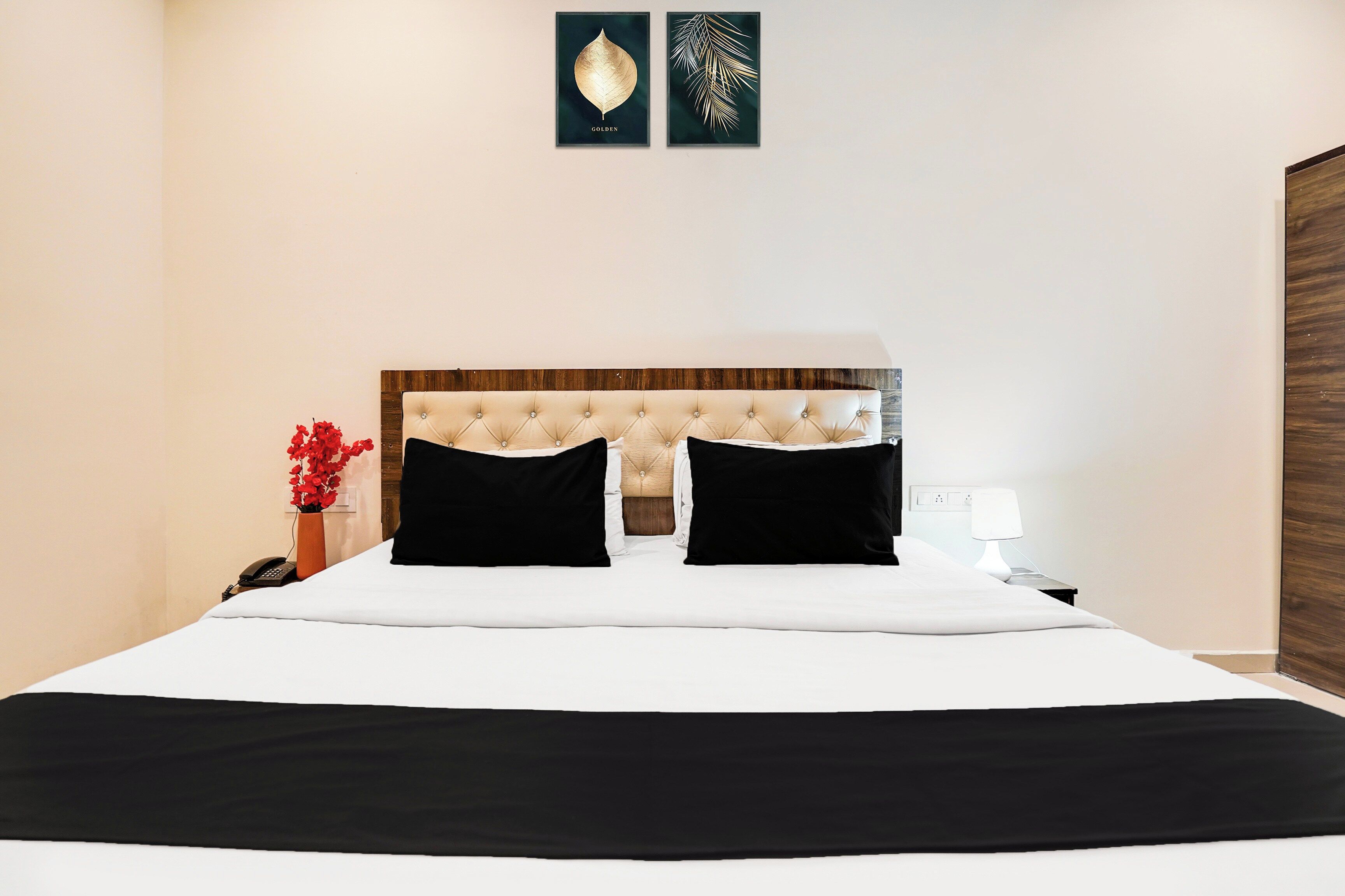 Una habitación de hotel con una cama grande, un cabecero de madera, una mesita de noche con lámpara y un jarrón con flores rojas.