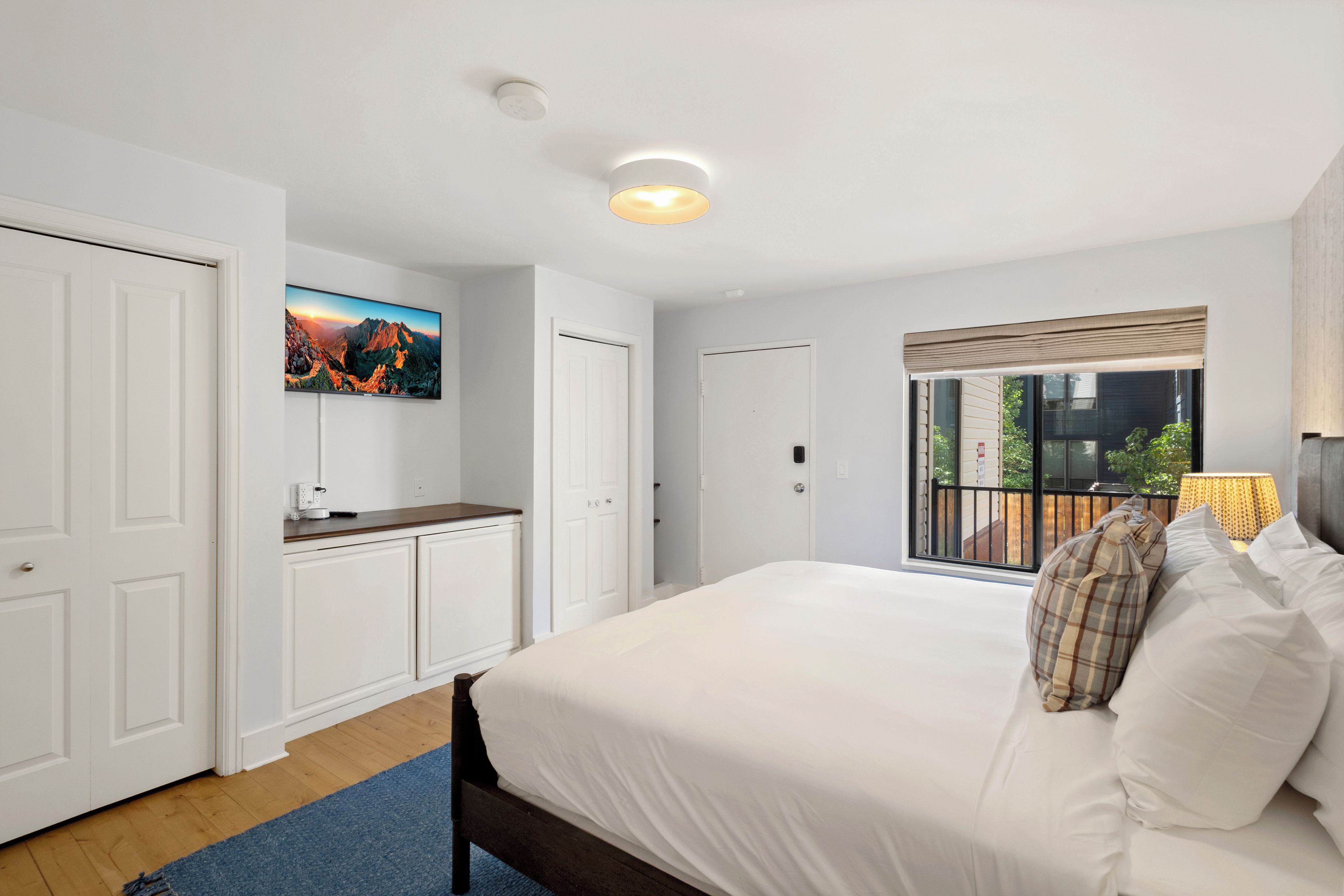 Un dormitorio con una cama grande, un televisor de pantalla plana montado en la pared, un ventanal con vistas al exterior y una lámpara en el techo.