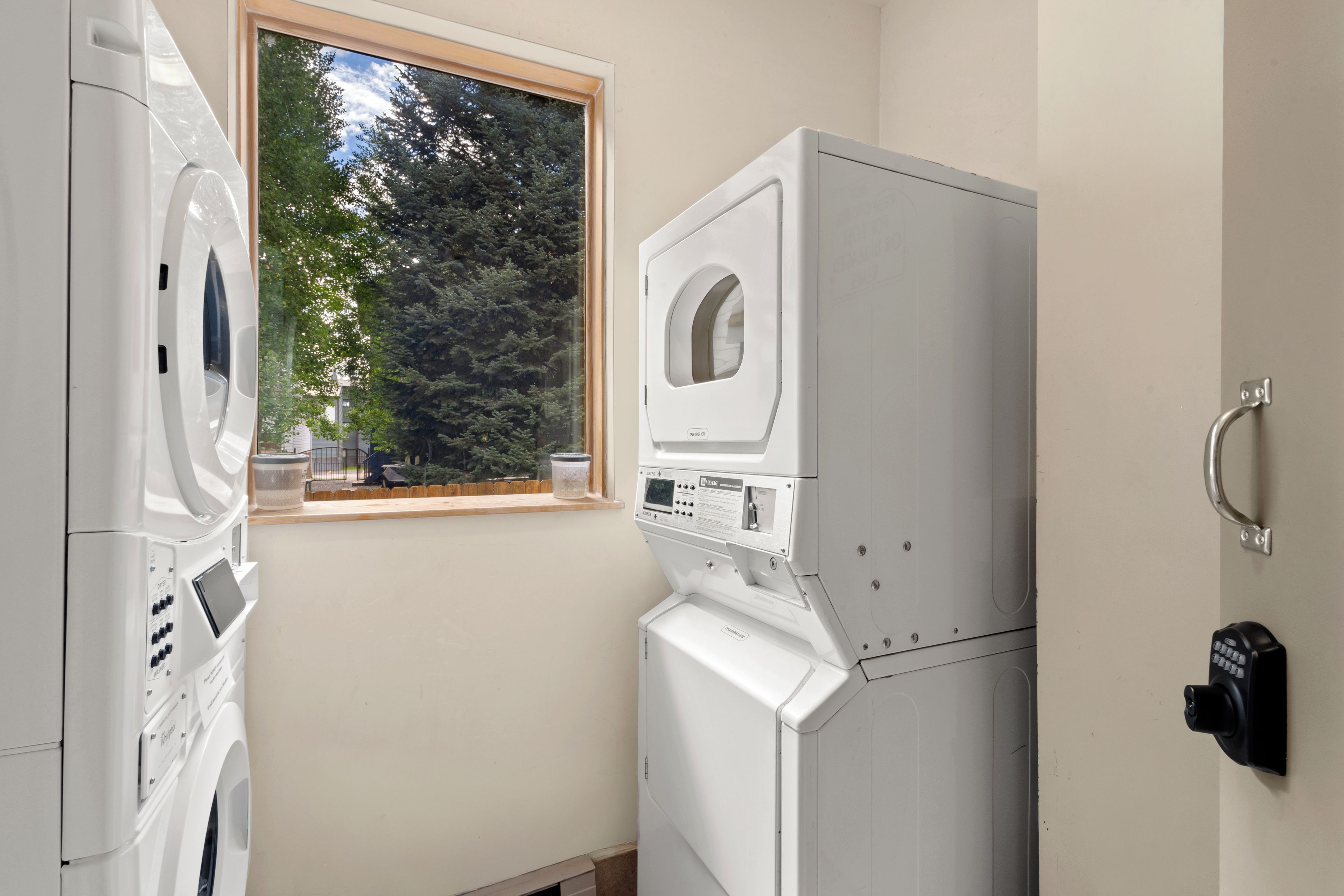 A laundry room with stacked washing machines and dryers, a window with a view of trees, and a wall-mounted security device.