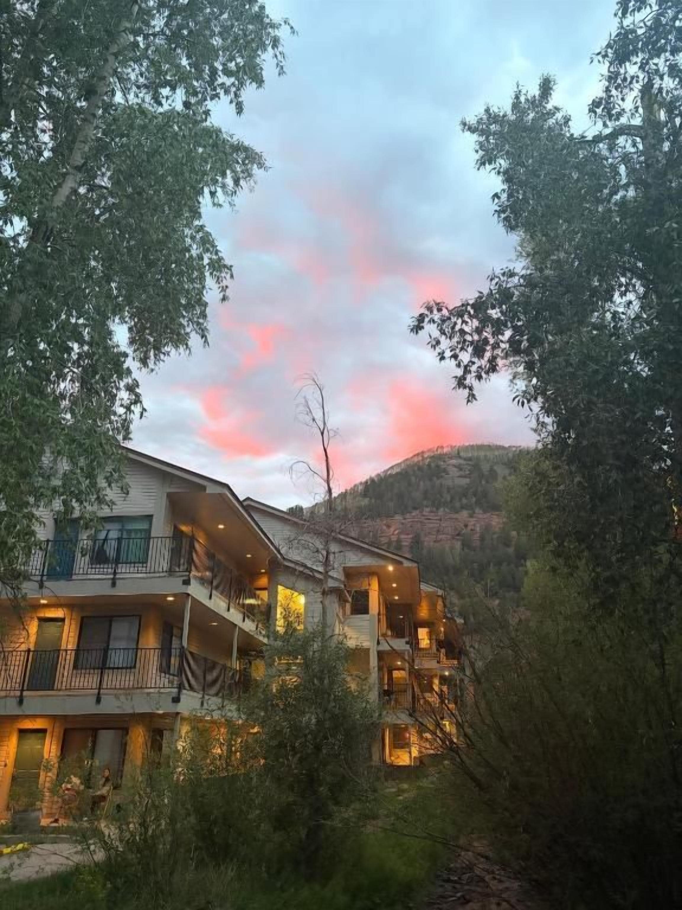 A multi-story building with balconies, surrounded by trees and a hilly landscape.
